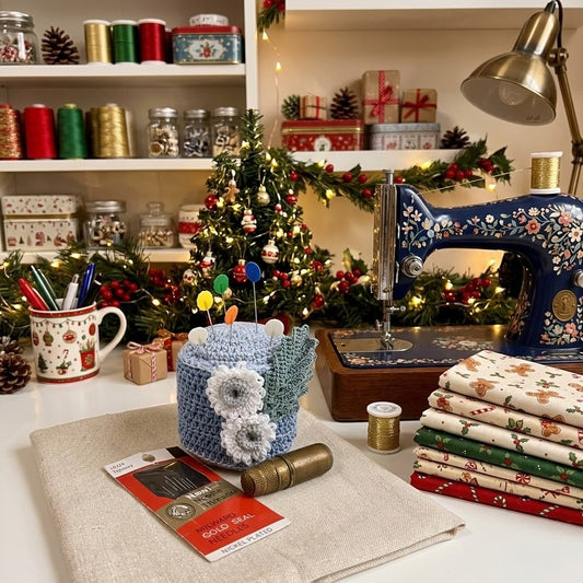 A cosy Christmas-themed sewing workspace with a blue floral vintage-style sewing machine, folded patterned fabric stacks, a crocheted pin holder, shelves of spools and jars, and a decorated Christmas tree in the background.