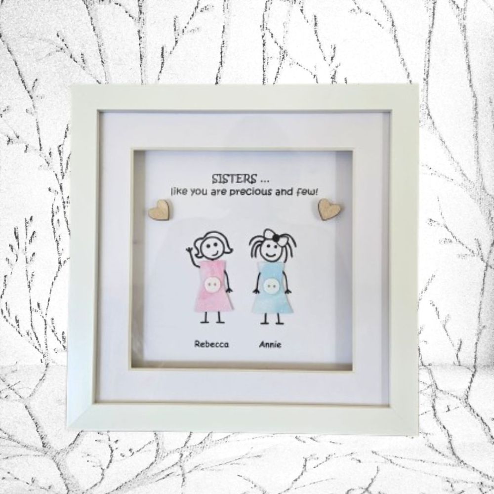 A square white shadow box frame containing a personalised children's print with the text 'SISTERS .