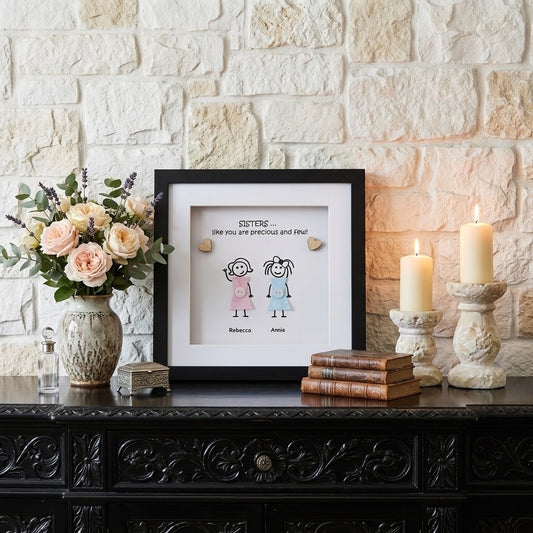 A framed custom illustration pebble art gift of two sisters displayed on a dark carved wooden console table, styled with a vase of pink and peach roses, candles, and books against a light stone wall.