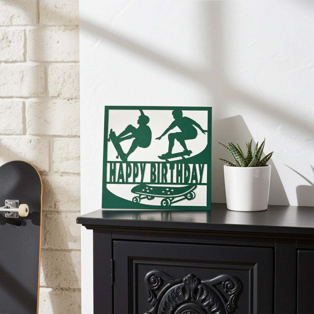 A square decorative card with a dark green and white skateboarder graphic and the words 'HAPPY BIRTHDAY' sits on a black cabinet in a styled room. A white brick wall, a potted succulent, and part of a skateboard are visible in the scene.