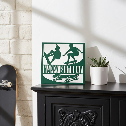 A square decorative card with a dark green and white skateboarder graphic and the words 'HAPPY BIRTHDAY' sits on a black cabinet in a styled room. A white brick wall, a potted succulent, and part of a skateboard are visible in the scene.