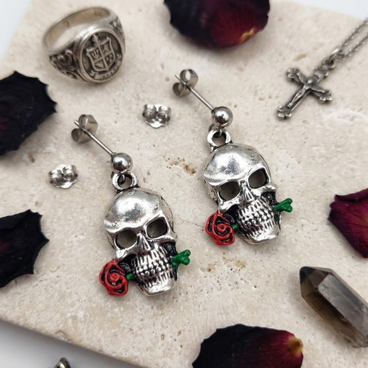 A pair of silver skull drop earrings with small red rose charms and green stems, displayed on a light stone surface with dried rose petals and jewelry props.
