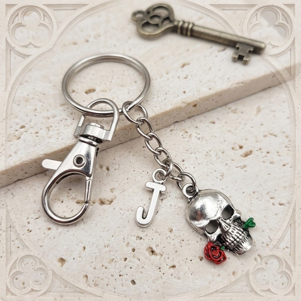 A silver-toned metal keychain featuring a lobster clasp, a letter 'J' charm, a skull charm, and a small red rose charm, displayed on a textured beige stone slab with a decorative key in the background.