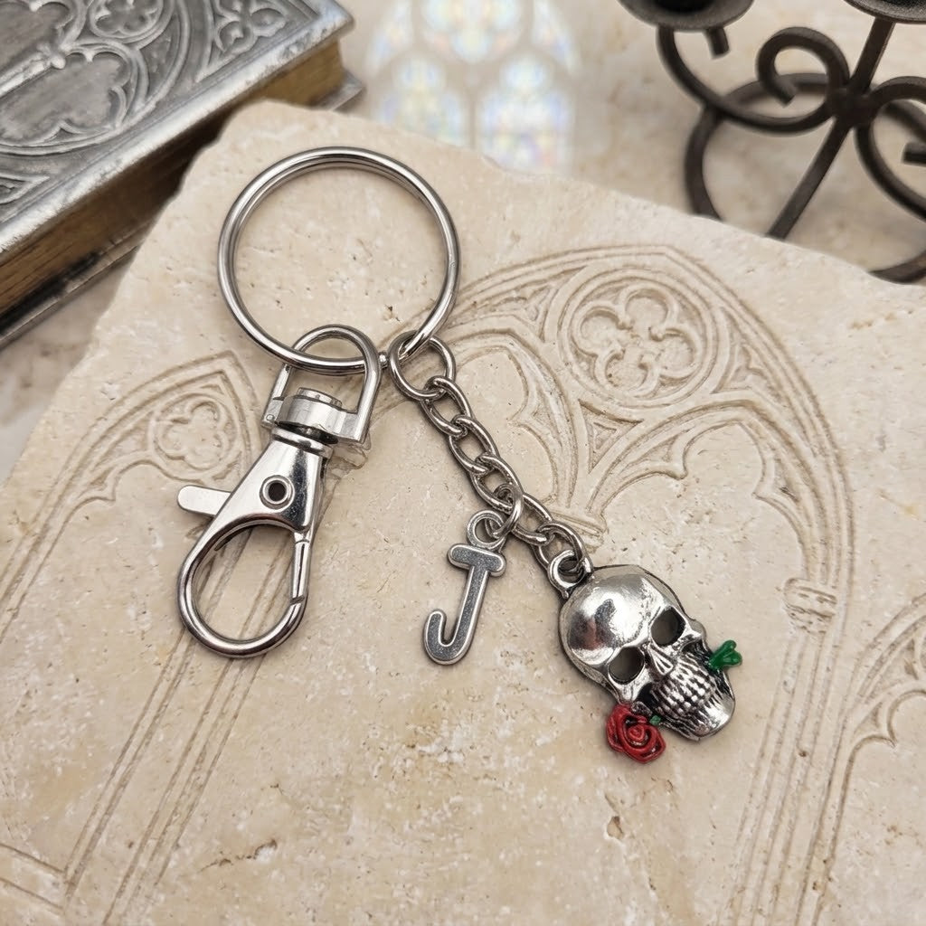 A silver-toned metal keychain featuring a lobster clasp, a letter 'J' charm, a skull charm with a small red rose, and a split key ring, displayed on a textured beige stone surface.