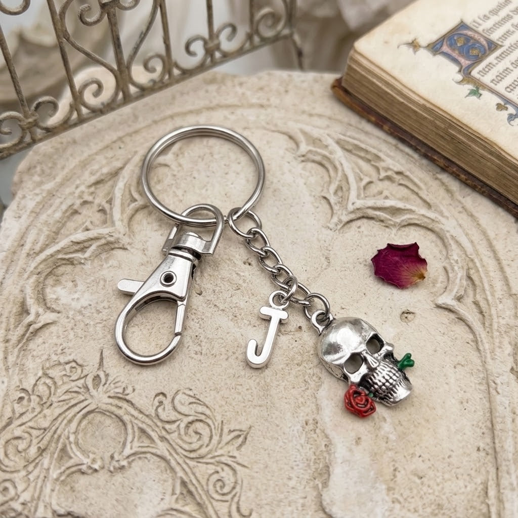 A silver-toned metal keychain with a lobster clasp, a hanging letter 'J' charm, a skull charm, and small red rose accents, displayed on a carved stone surface with an old book and metal gate in the background.