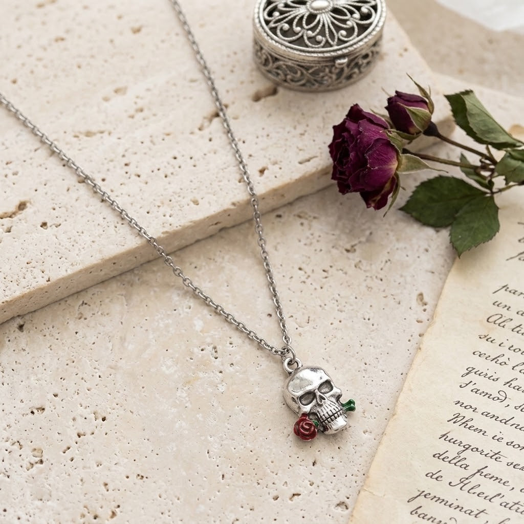 A silver skull pendant necklace with a small red rose and green leaf charm, displayed on a textured beige stone slab with a vintage book page and a dried rose bud as props.