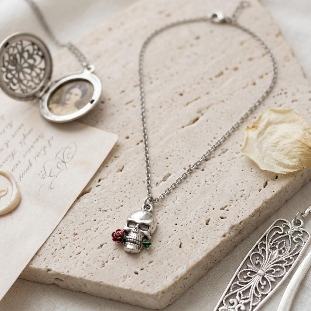 A silver skull pendant necklace with a small red rose and green leaves accent, displayed on a textured beige stone slab with vintage props including a locket, dried flower, and letter.