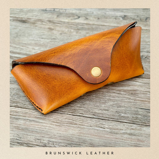 Brown leather sunglasses case on a wooden surface with 'Brunswick Leather' branding.