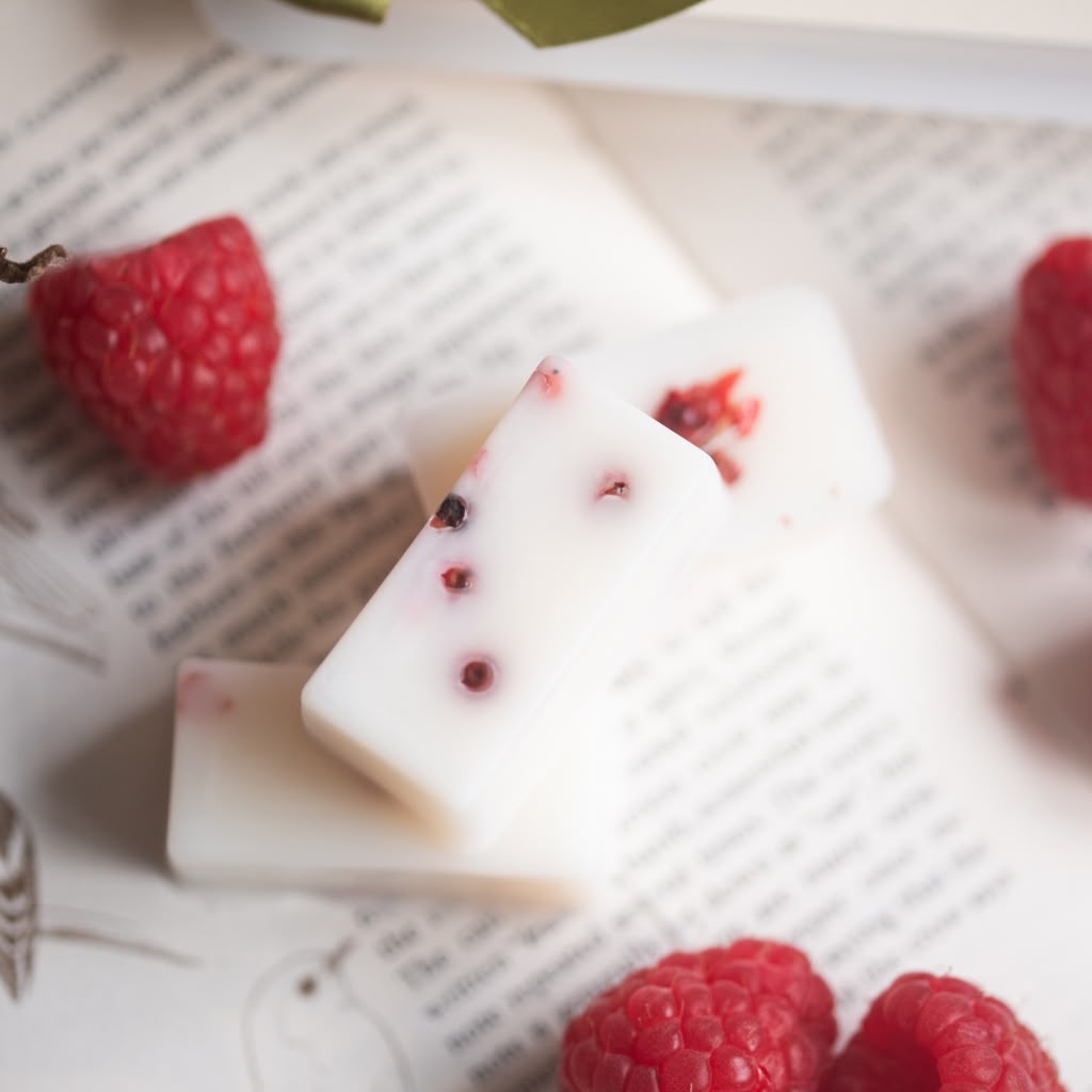 White rectangular wax melts with red berry inclusions, arranged on an open book surrounded by fresh raspberries.