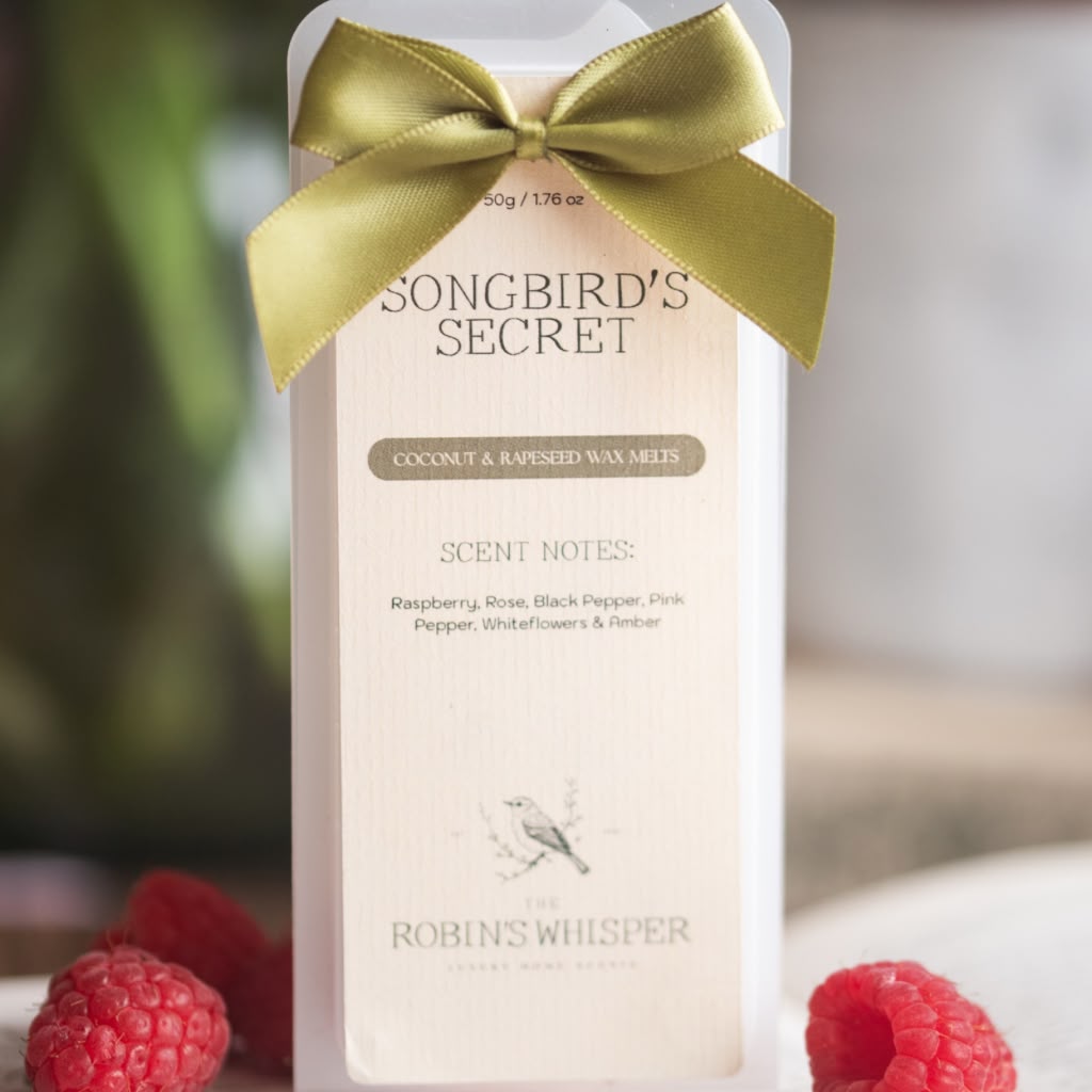 A boxed fragrance product labelled 'SONGBIRD'S SECRET' with a pale blush pink package, a gold ribbon bow, and raspberries placed around it on a softly blurred background.