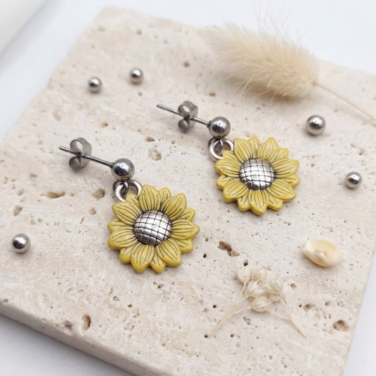 A pair of yellow sunflower dangle earrings with textured petals and silver-toned metal posts, displayed on a beige stone tile with dried grass and small metallic beads.