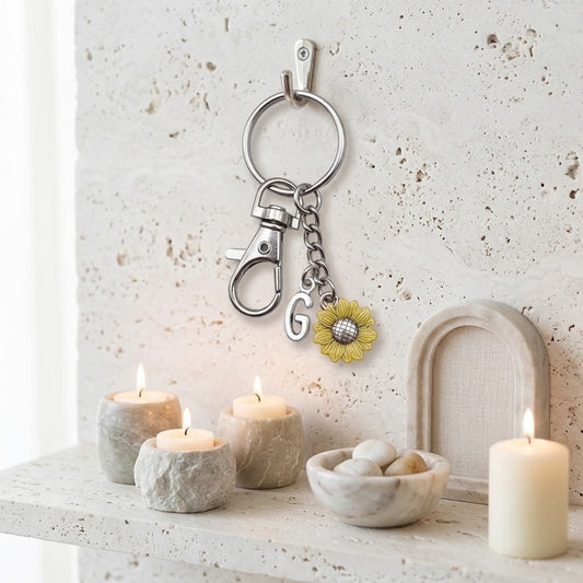 A silver metal keychain with a lobster clasp, a hanging chain, a letter G charm, and a yellow sunflower charm, displayed on a textured wall above a shelf with lit candles and decorative bowls.
