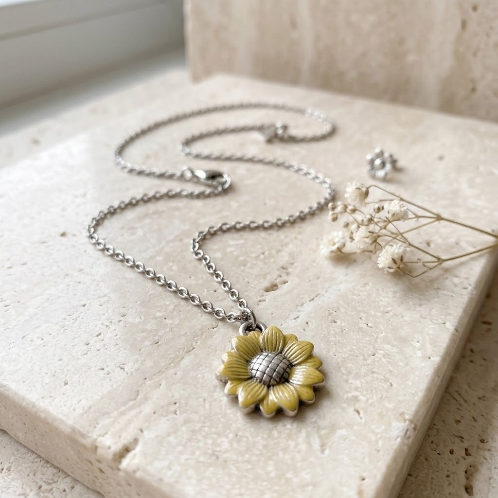 A silver chain necklace with a yellow and silver sunflower pendant, styled on a beige stone slab with dried baby's breath flowers.
