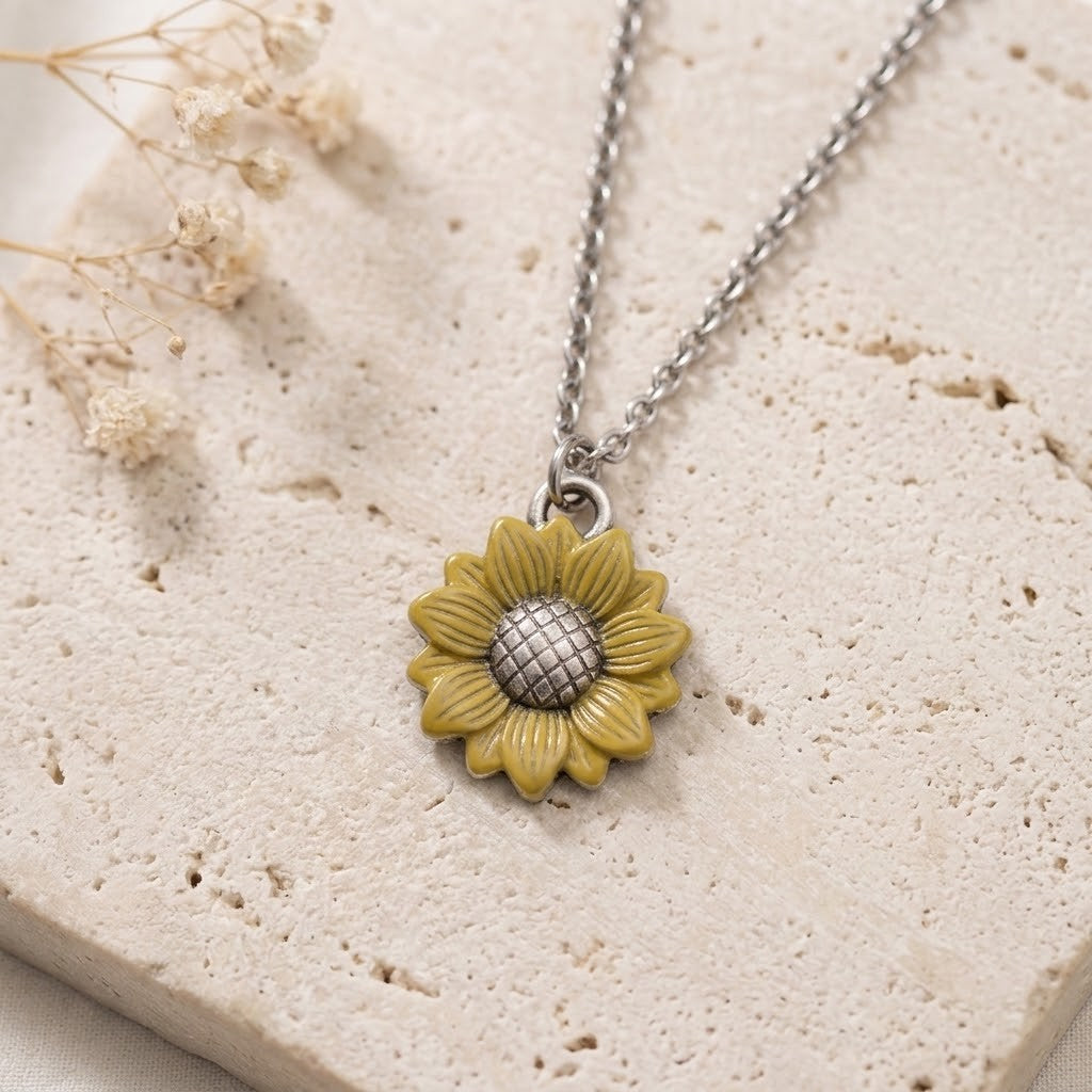 A close-up of a sunflower pendant necklace on a silver chain, resting on a textured beige stone surface with dried flowers in the corner.