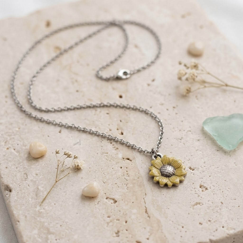 A silver chain necklace with a yellow and silver flower pendant, styled on a textured beige stone slab with dried flowers and sea glass.