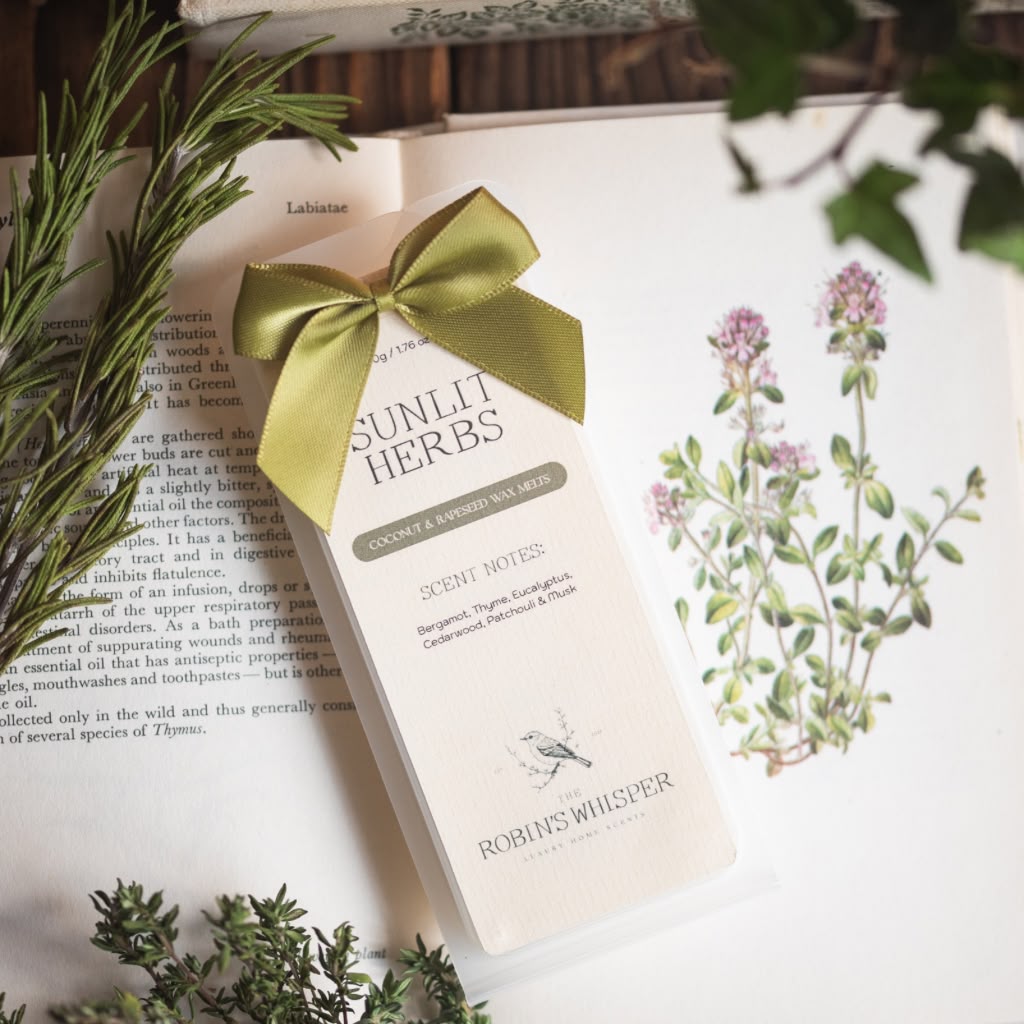 A pack of wax melts from Robins Whisper, tied with a green satin ribbon, resting on an open book surrounded by botanical illustrations and green sprigs.