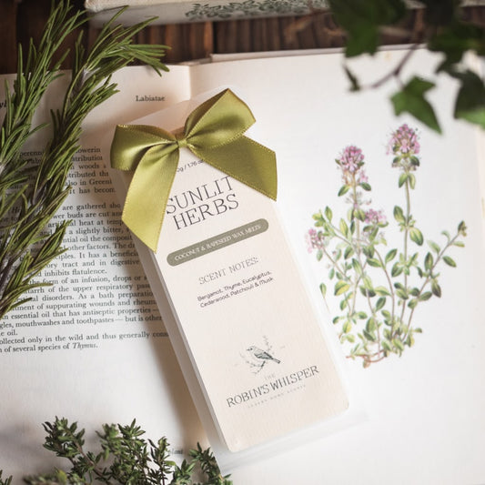 A pack of wax melts from Robins Whisper, tied with a green satin ribbon, resting on an open book surrounded by botanical illustrations and green sprigs.