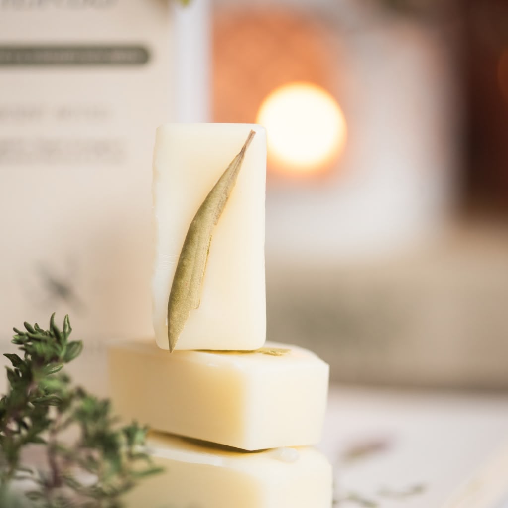 Three rectangular bars of creamy off-white wax melts stacked vertically, with a dried green leaf pressed onto the top bar. The scene is styled with green sprigs in the foreground and a warm, softly blurred background with a glowing light source.