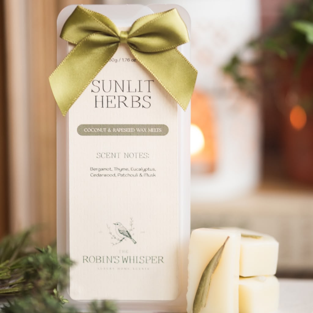 A boxed wax melt product labelled 'Sunlit Herbs' tied with a gold ribbon, shown in a softly blurred indoor setting with lit candles and greenery in the background. Two cream-coloured wax melts with gold ribbon accents sit in front.