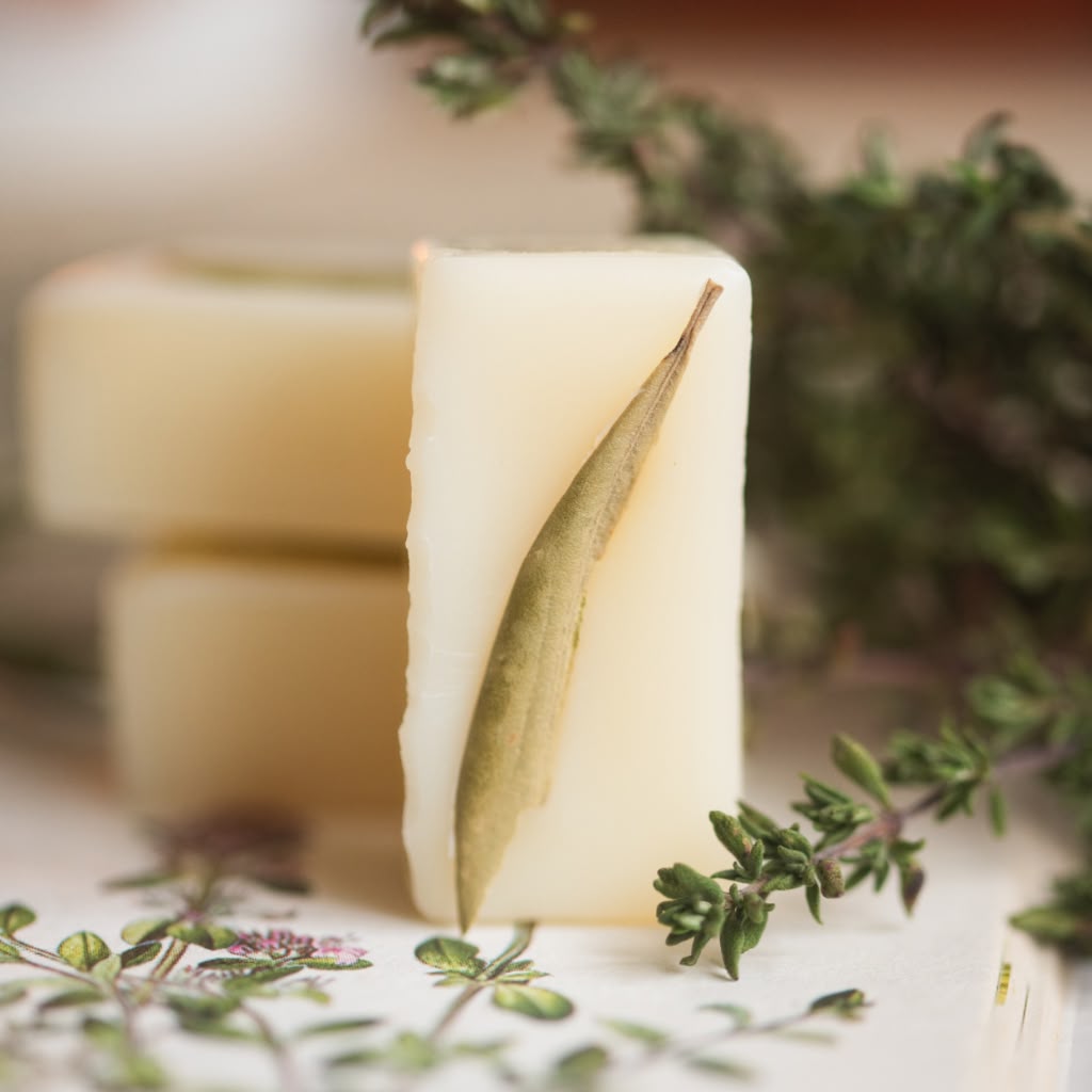 A rectangular bar of pale cream wax melt stands upright on a white surface, with a dried green leaf pressed onto one side. Two more soap bars are softly blurred in the background, surrounded by green herb stems and leaves.