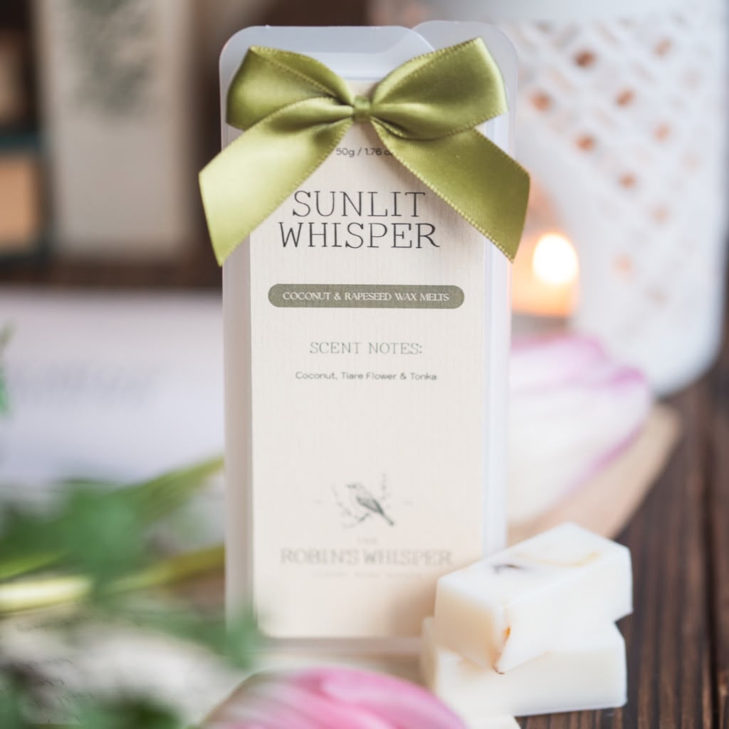 A clear plastic package of Sunlit Whisper coconut and rapeseed scented wax melts tied with an olive green satin bow, shown on a wooden surface with a lit candle and wax pieces in the background.