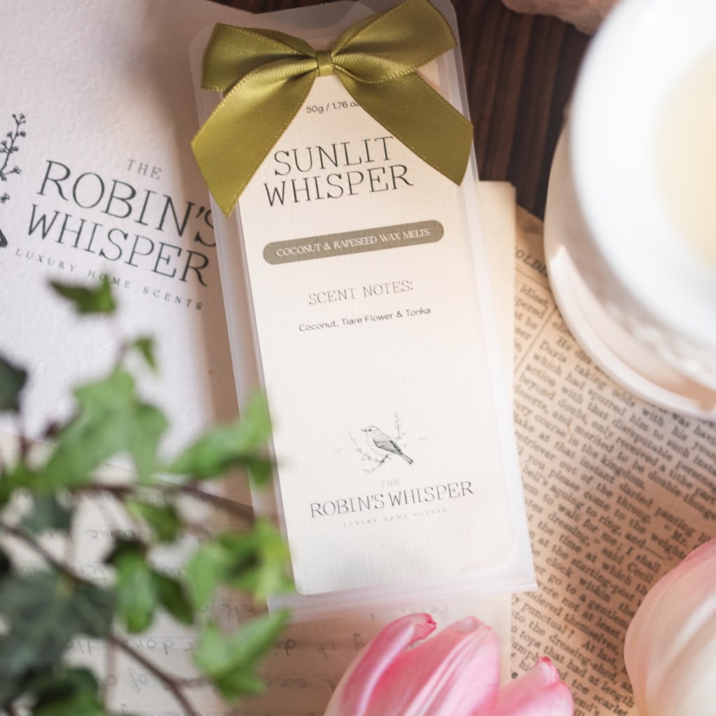 A boxed wax melt product labelled 'Sunlit Whisper' by Robin's Whisper, wrapped in a cream paper sleeve with a gold ribbon bow, styled on top of printed paper with greenery and pink flowers.