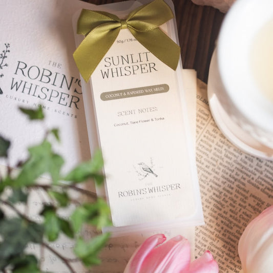 A boxed wax melt product labelled 'Sunlit Whisper' by Robin's Whisper, wrapped in a cream paper sleeve with a gold ribbon bow, styled on top of printed paper with greenery and pink flowers.