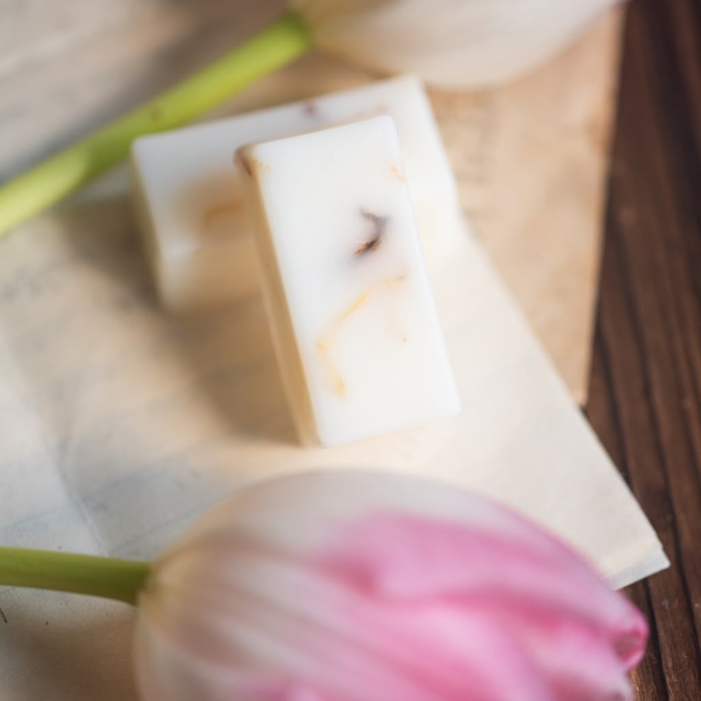 Two rectangular bars of handmade wax melts with white and cream marbled patterns, resting on a piece of parchment paper with a pink tulip in the foreground and a wooden surface in the background.