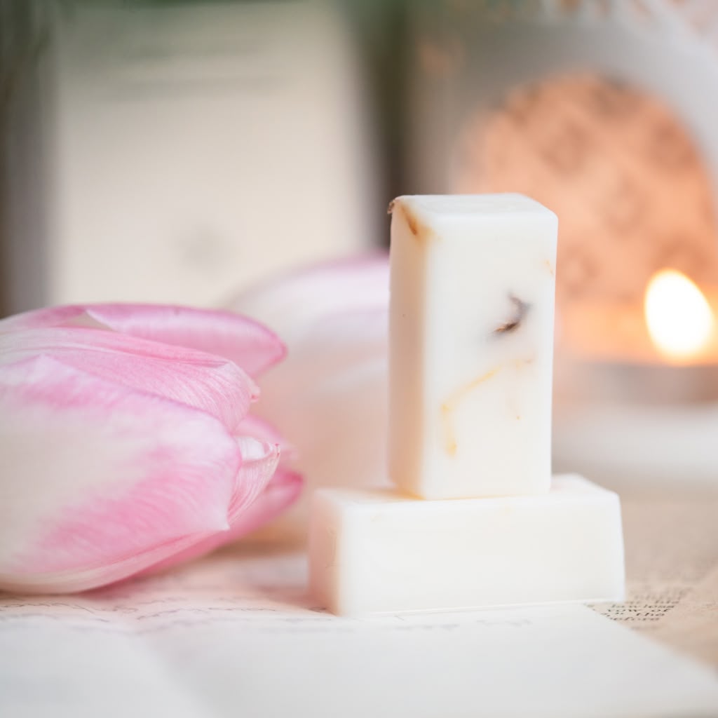 A small rectangular bar of wax melt with subtle tan and dark inclusions, displayed on a matching white soap block next to a pink tulip flower against a softly blurred background with a warm light.