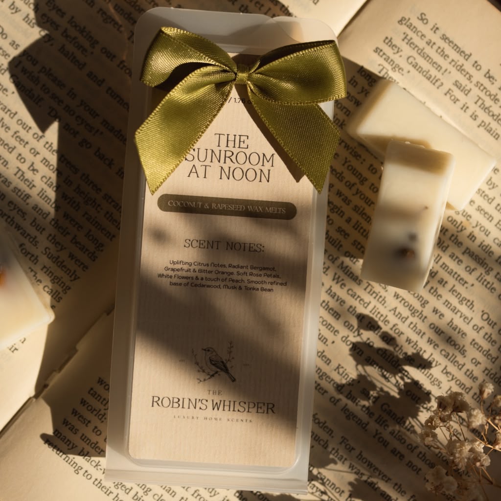 A boxed scented wax melt from Robin's Whisper is displayed on an open book, tied with a gold satin bow. The packaging is cream-colored with black text reading 'THE SUNROOM AT NOON' and 'SCENT MOIST'.