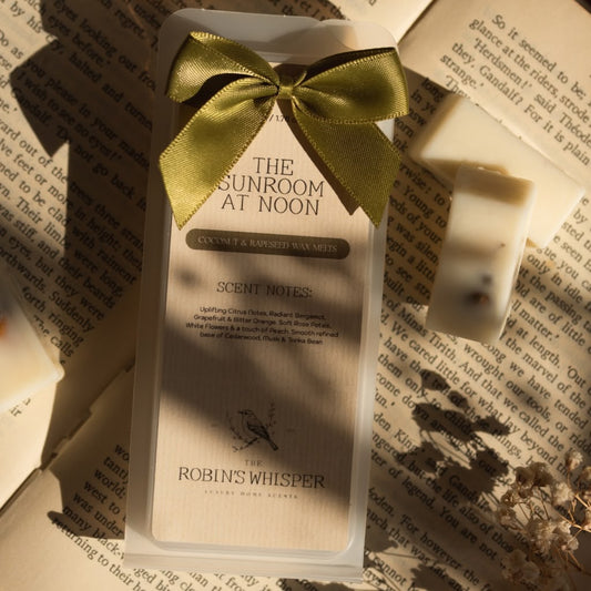A boxed scented wax melt from Robin's Whisper is displayed on an open book, tied with a gold satin bow. The packaging is cream-colored with black text reading 'THE SUNROOM AT NOON' and 'SCENT MOIST'.
