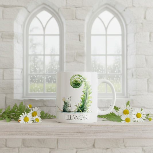 White mug with green design and 'Eleanor' text on a white surface with flowers, against a white stone wall background.