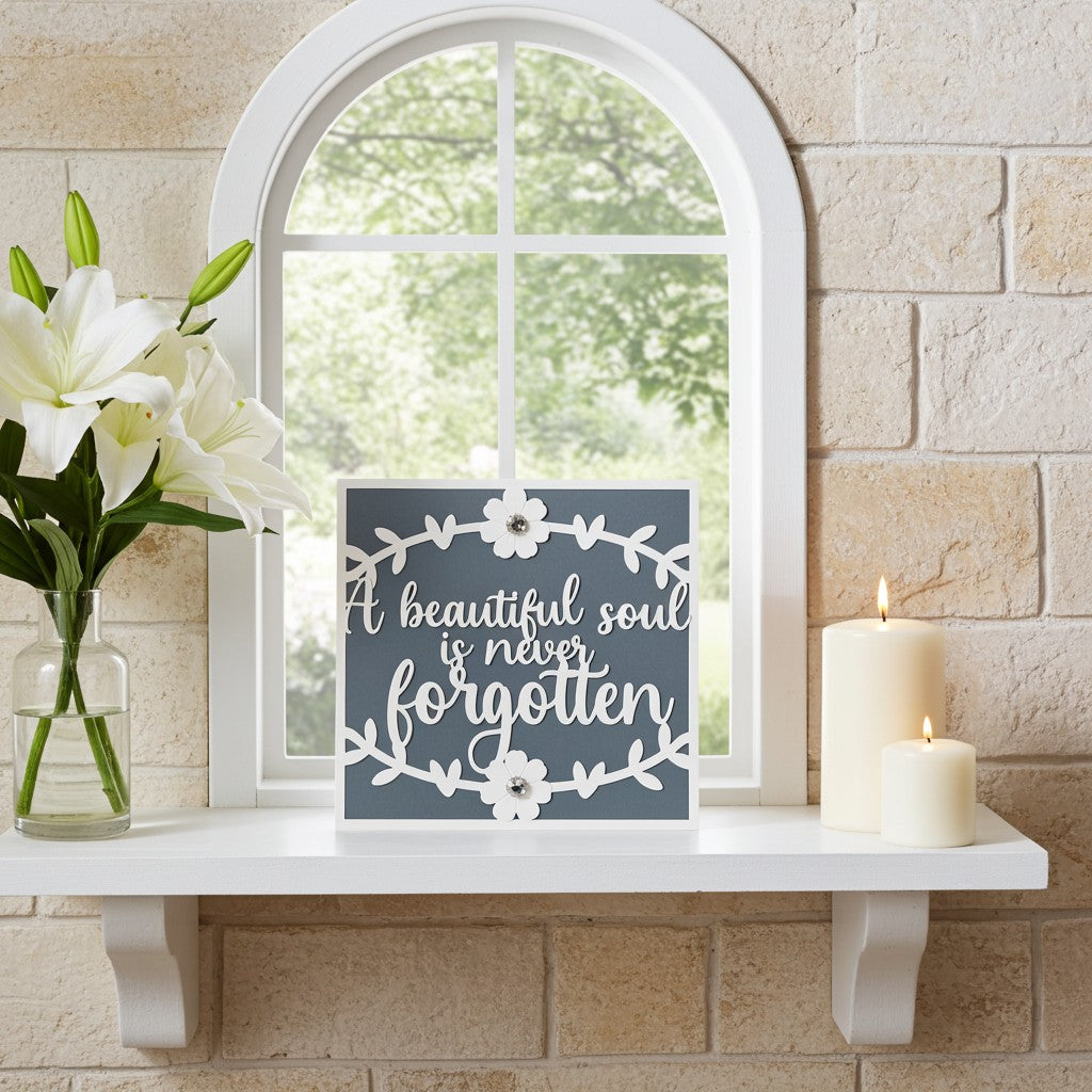 A square decorative sympathy card with the phrase “A beautiful soul is never forgotten” in white script on a dark grey background, styled on a white shelf next to white lilies in a glass vase and two lit white pillar candles against a beige stone wall and arched window.