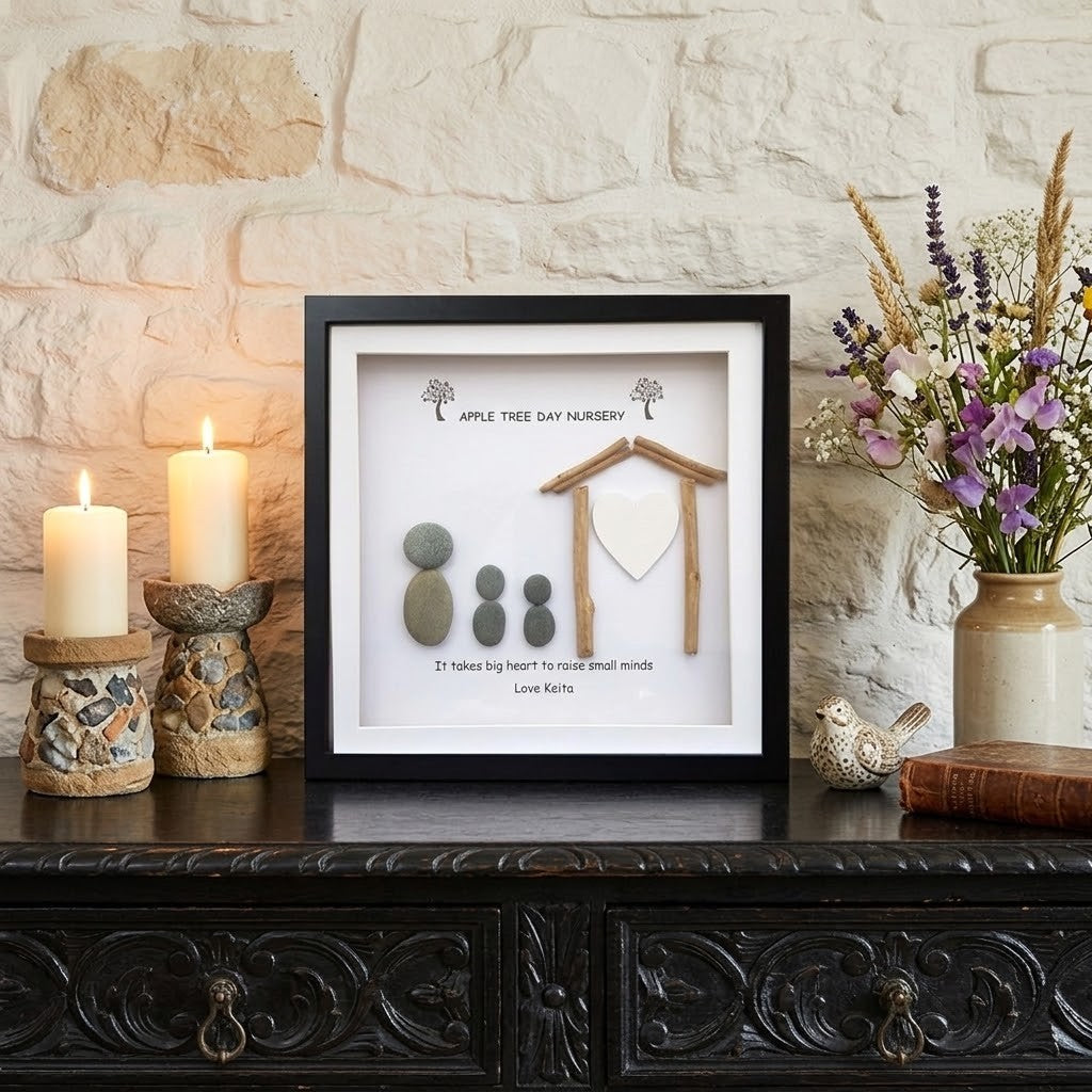 A framed shadow box display on a dark carved wooden table against a light stone wall. The display includes small stones, a wooden house shape, and printed text reading 'Apple Tree Day Nursery' 
