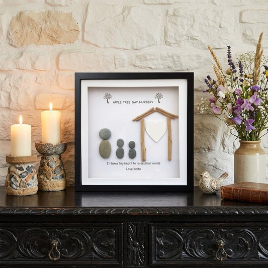 A framed shadow box display on a dark carved wooden table against a light stone wall. The display includes small stones, a wooden house shape, and printed text reading 'Apple Tree Day Nursery'