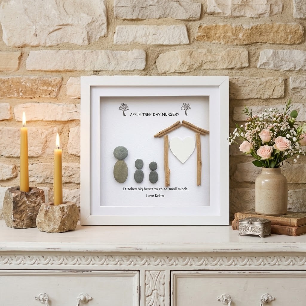 A framed personalised teacher gift display on a white console table against a stone wall. The white frame contains a small wooden house, a white heart, three grey stones, and printed text reading 'Apple Tree Day Nursery' and 'It takes big hearts to root small minds'