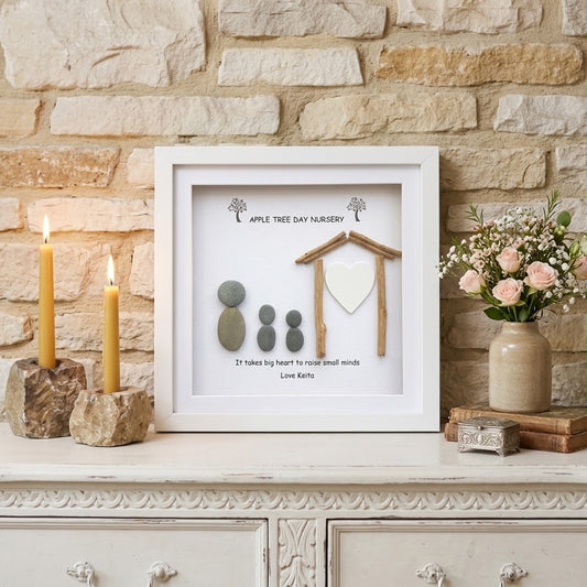 A framed personalised teacher gift display on a white console table against a stone wall. The white frame contains a small wooden house, a white heart, three grey stones, and printed text reading 'Apple Tree Day Nursery' and 'It takes big hearts to root small minds'