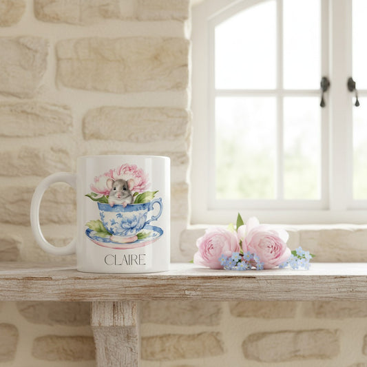 Mug with a design of a mouse in a teacup and flowers, placed on a wooden surface with a stone wall and window in the background.