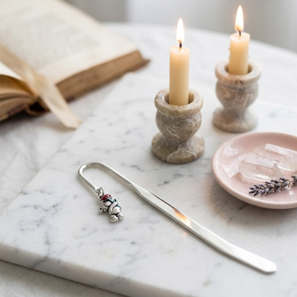 A silver metal bookmark with a small teddy bear charm and red berries, styled on a white marble tray alongside two lit candles in stone holders, an open book, and a pink dish.