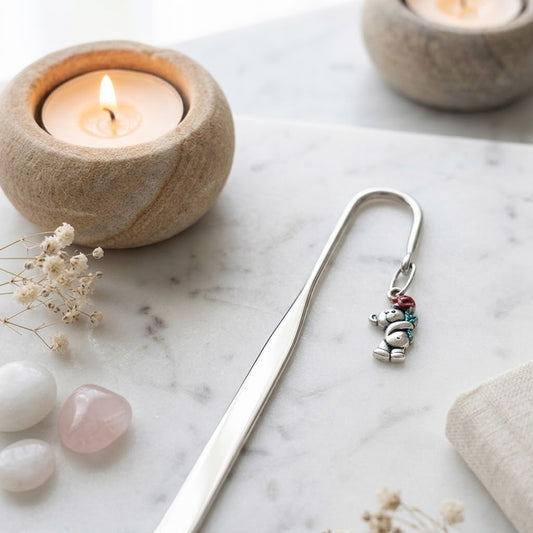 A silver metal bookmark shaped like a small snowman with colourful red, blue, and green accents, resting on a white marble surface next to a lit candle holder and dried flowers.