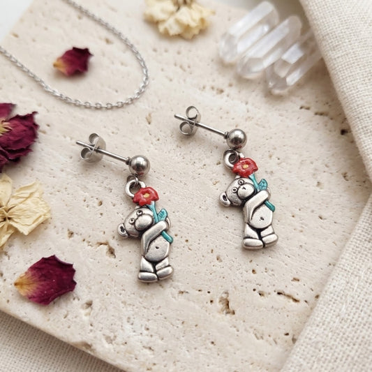 A pair of silver dangle earrings featuring a teddy bear holding a red rose, styled on a textured stone surface with dried flowers, crystal points, and a silver necklace.