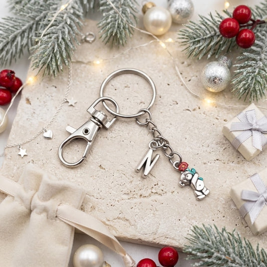 A silver-toned metal keychain with a lobster clasp, a split key ring, a hanging letter 'N' charm, and a small teddy bear charm with red and green accents, styled on a stone surface with Christmas decorations.