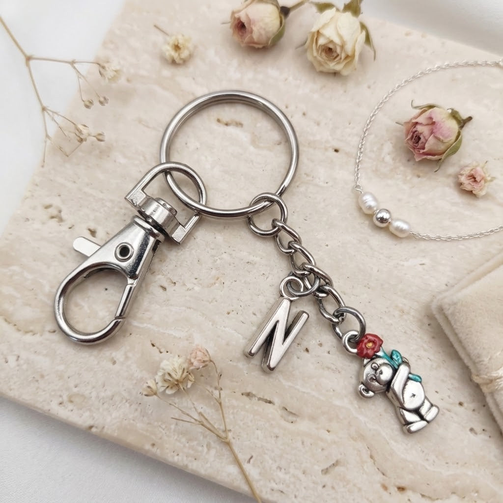 A silver-toned metal keychain with a lobster clasp, a letter 'N' charm, and a small bear charm with red and teal beads, styled on a beige stone slab with dried flowers.