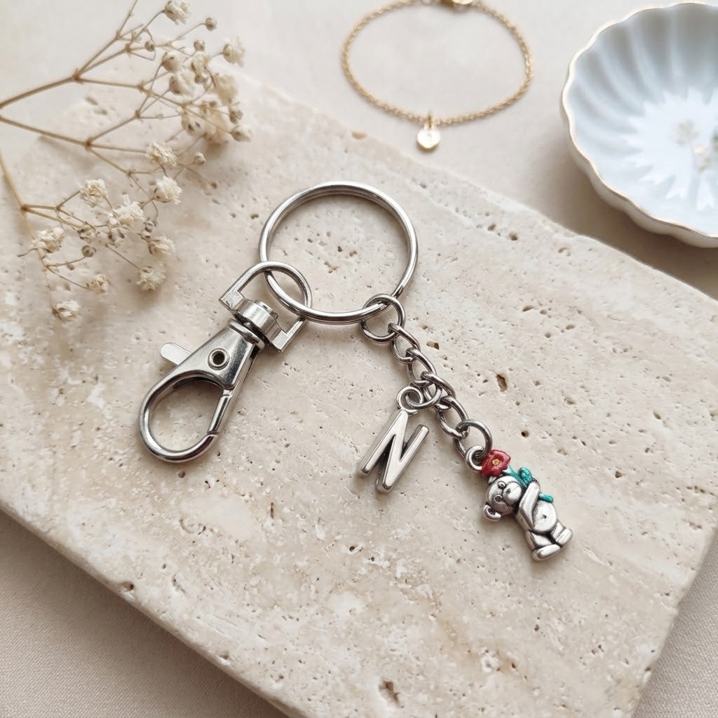 A silver-toned keychain with a lobster clasp, a hanging letter charm, and a small bear charm holding a red and green balloon, styled on a beige stone slab with dried flowers and a small dish in the background.