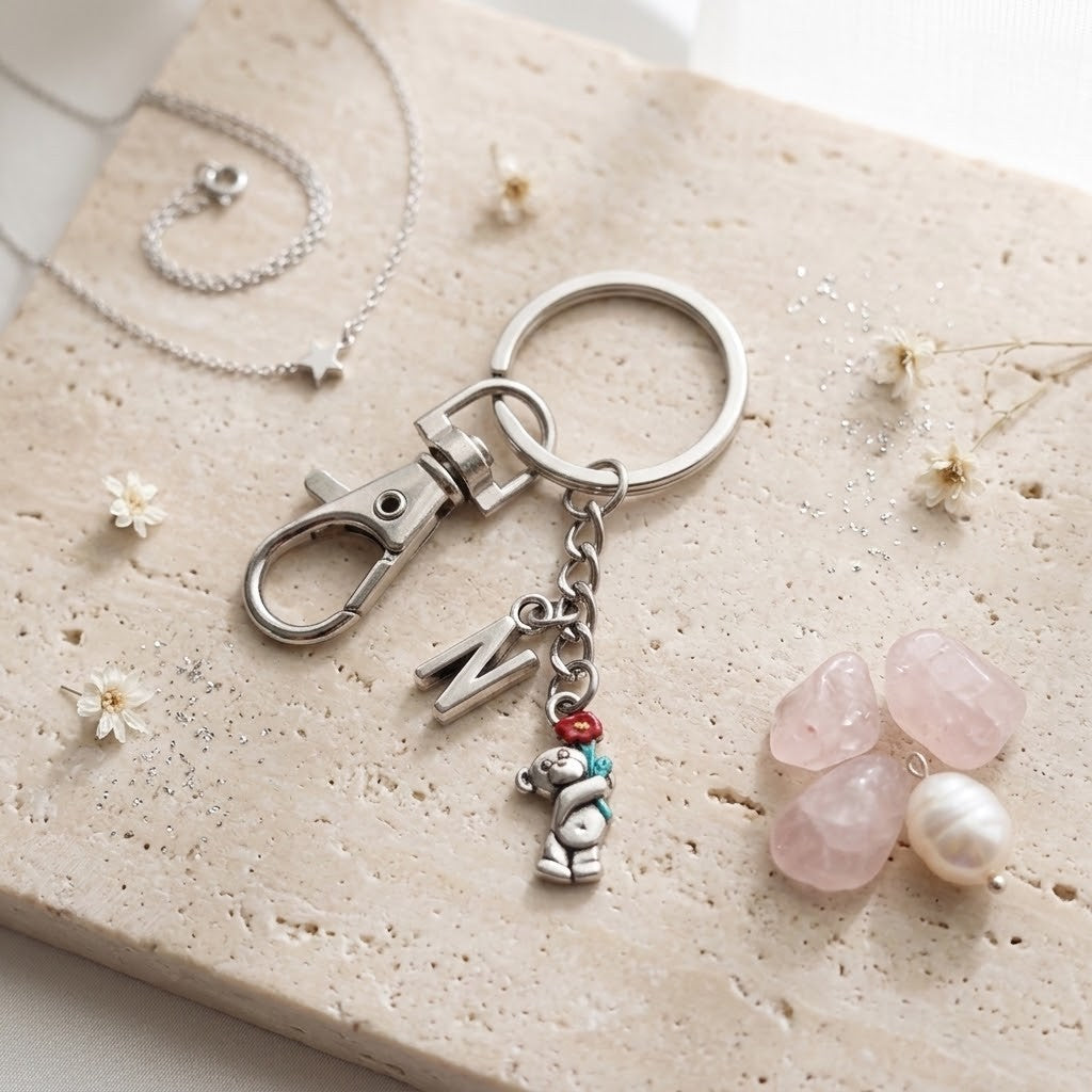 A silver-toned keychain with a lobster clasp, a split key ring, and dangling charms including a letter 'M', a small bear charm with turquoise and red accents, and a keyring, styled on a beige stone slab with pink stones and dried flowers.