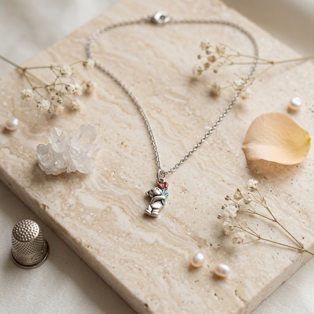 A silver-toned chain necklace with a small bear pendant resting on a beige stone slab, surrounded by dried flowers, pearls, a crystal cluster, and a metal thimble.