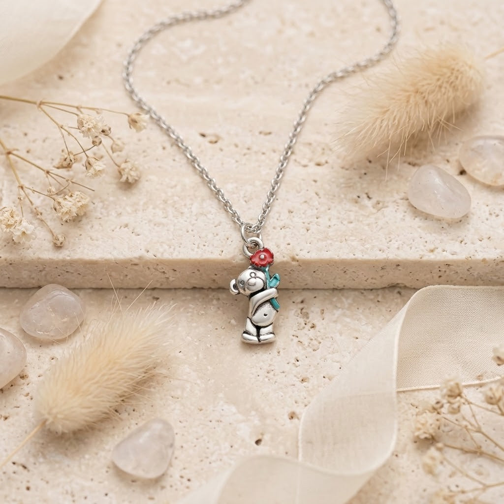 A silver pendant necklace featuring a teddy bear character holding a red flower and a green bow, displayed on a textured beige stone surface with dried grass and clear stones.
