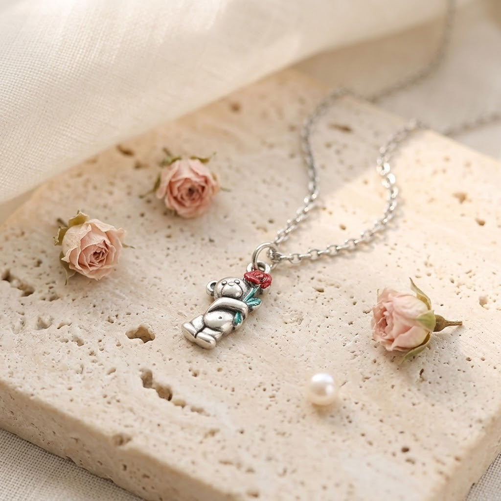 A silver-toned necklace with a small bear charm holding a red heart and a blue-green gem, displayed on a textured beige stone slab with pink roses and a pearl.