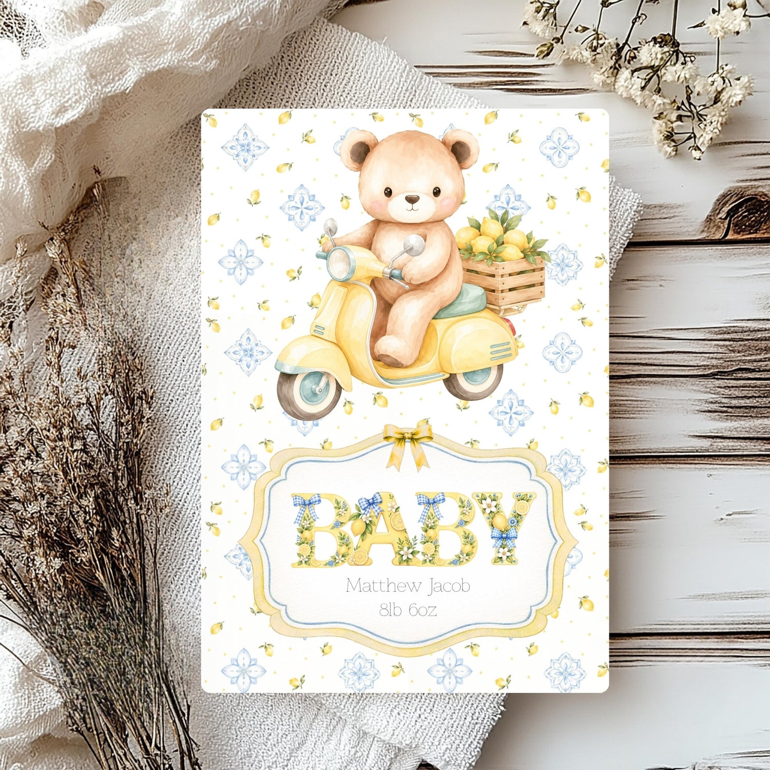 A personalised baby card featuring a cute teddy bear on a yellow scooter, styled on a rustic wooden surface with neutral fabric and dried flowers.