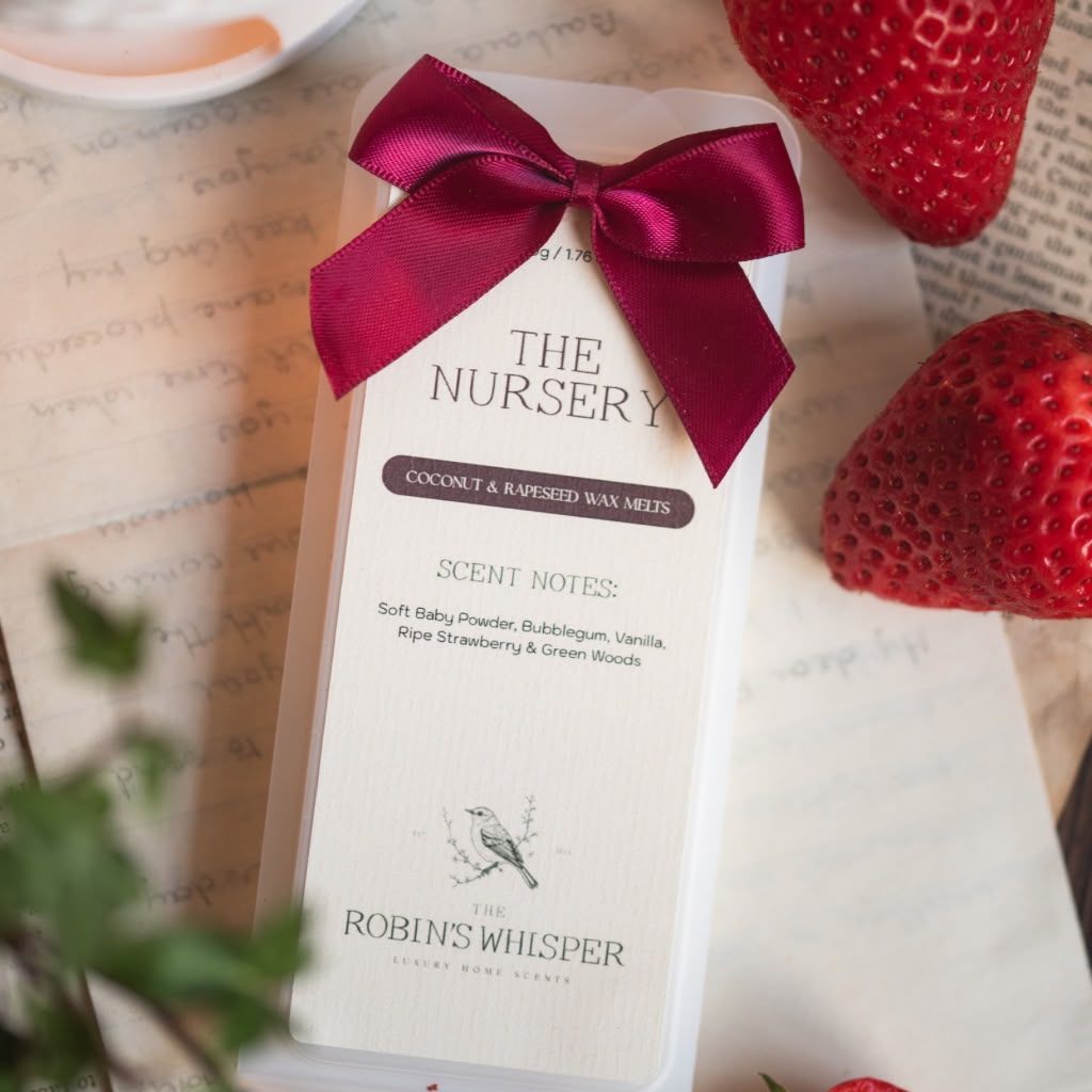 A wax melt package for The Nursery by The Robin's Whisper, tied with a dark red satin bow, displayed on a vintage book page with strawberries as props.
